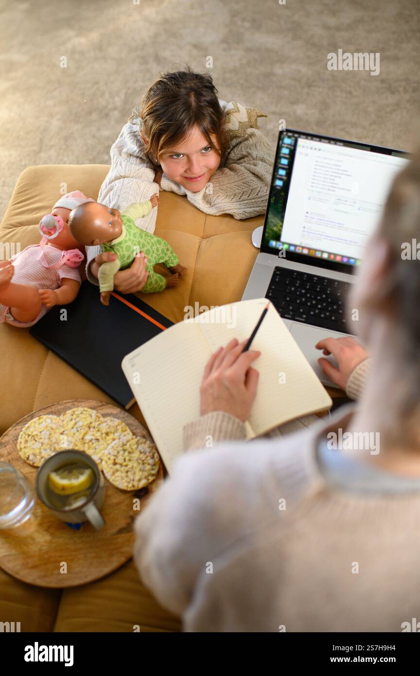 Una madre lavora sul suo portatile a casa mentre sua figlia aspetta pazientemente per giocare con le bambole insieme. Foto Stock