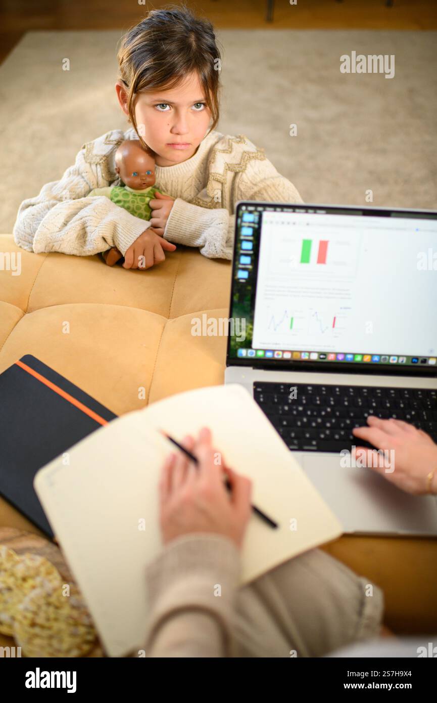 Una madre lavora sul suo portatile a casa mentre sua figlia aspetta pazientemente per giocare con le bambole insieme. Foto Stock