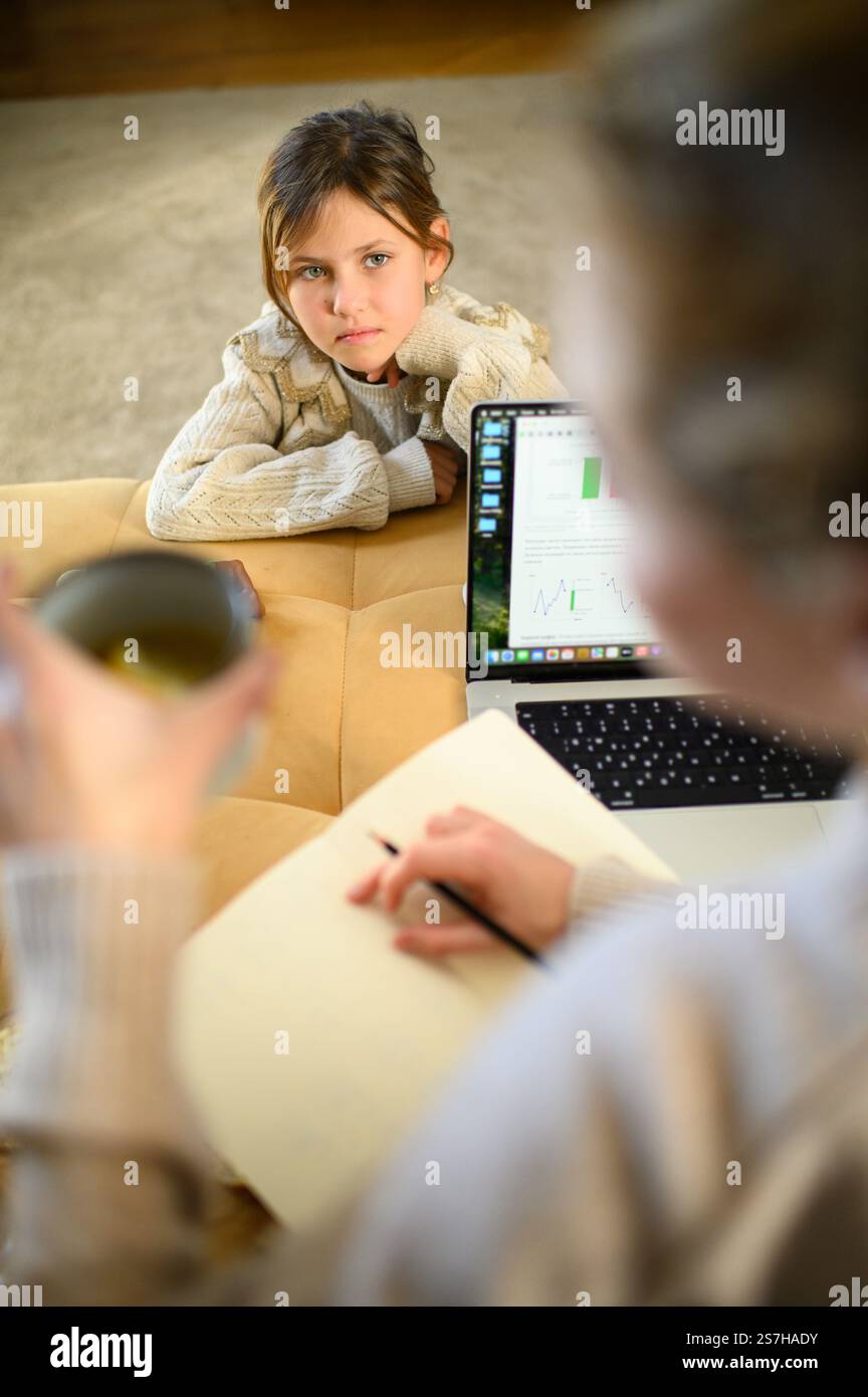 Una madre lavora sul suo portatile a casa mentre sua figlia aspetta pazientemente per giocare con le bambole insieme. Foto Stock