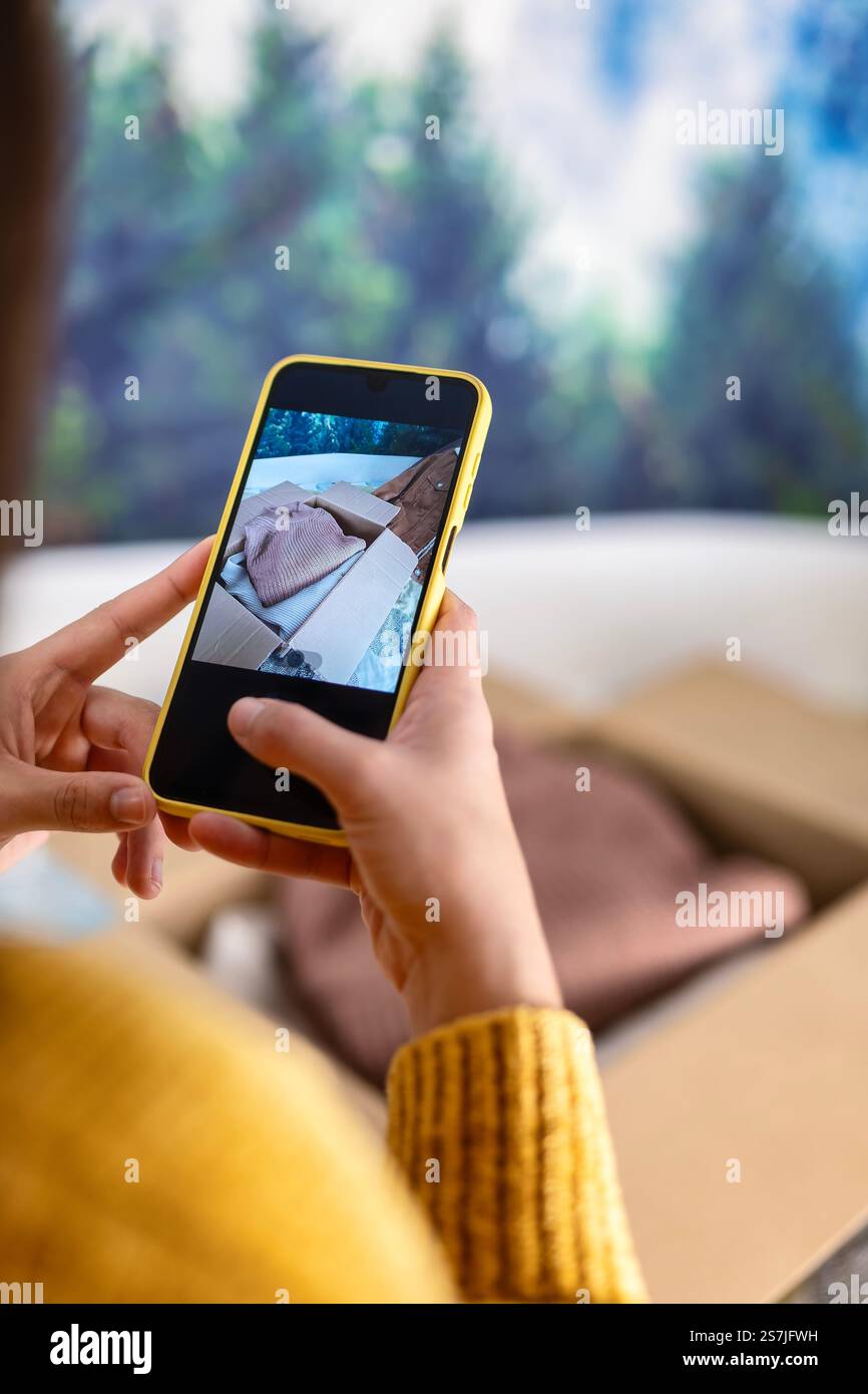 Donna che cattura immagini di abbigliamento da vendere con uno smartphone, in un ambiente domestico con pacchi pronti per la spedizione, seconda mano, commercio digitale e AN Foto Stock