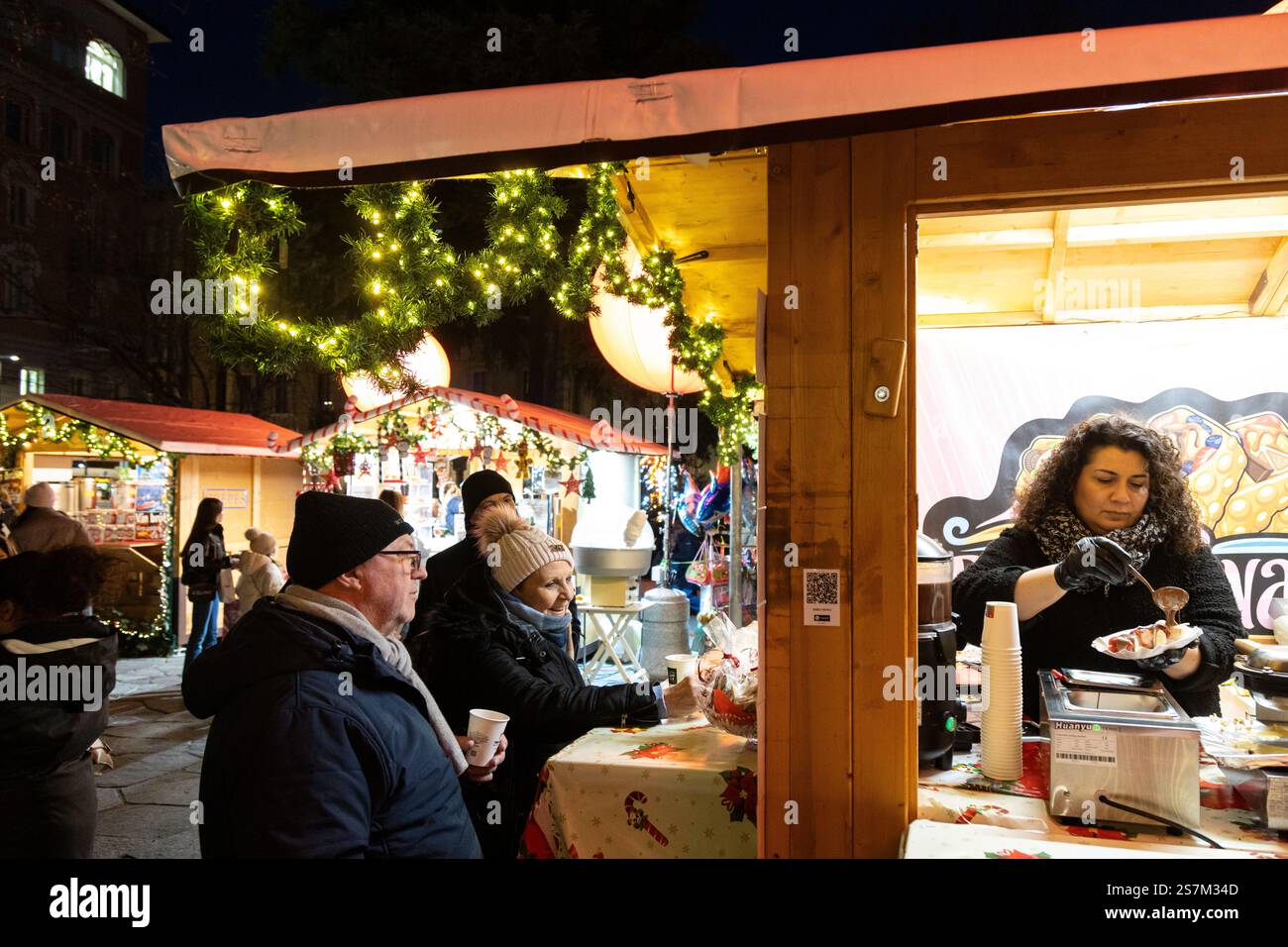 Lugano, Svizzera - 23 dicembre 2024: Visitatori in una banchina di mercatini di Natale che si godono le prelibatezze natalizie di notte con decorazioni e luci Foto Stock