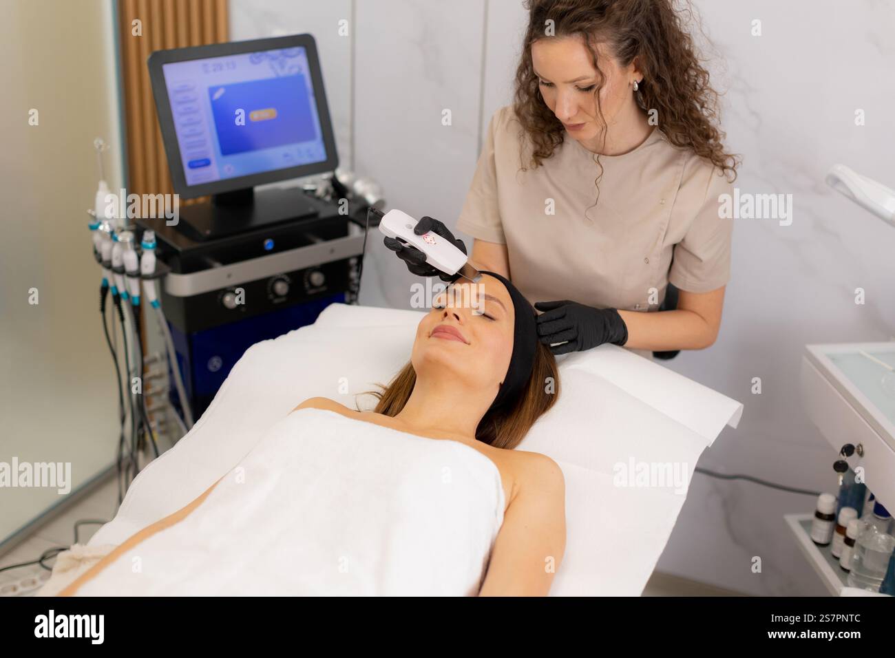 Un terapista esperto utilizza delicatamente un massaggiatore facciale a ultrasuoni sulla fronte del cliente, migliorando il relax e la cura della pelle in un tranquillo ambiente termale Foto Stock