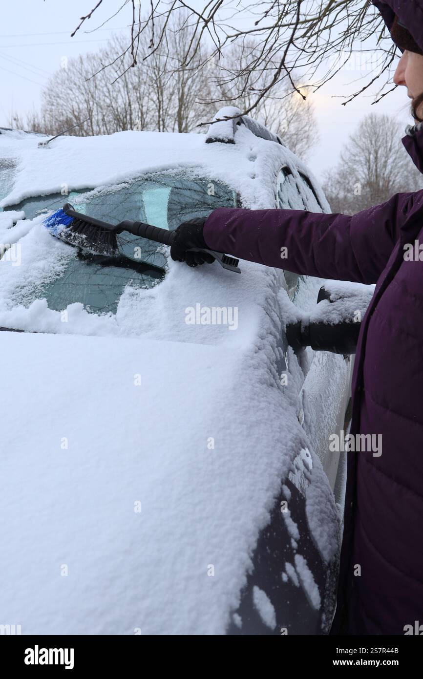 Vista laterale di una giovane donna con abiti caldi che puliscono la neve da un'auto con uno speciale orientamento verticale della spazzola Foto Stock