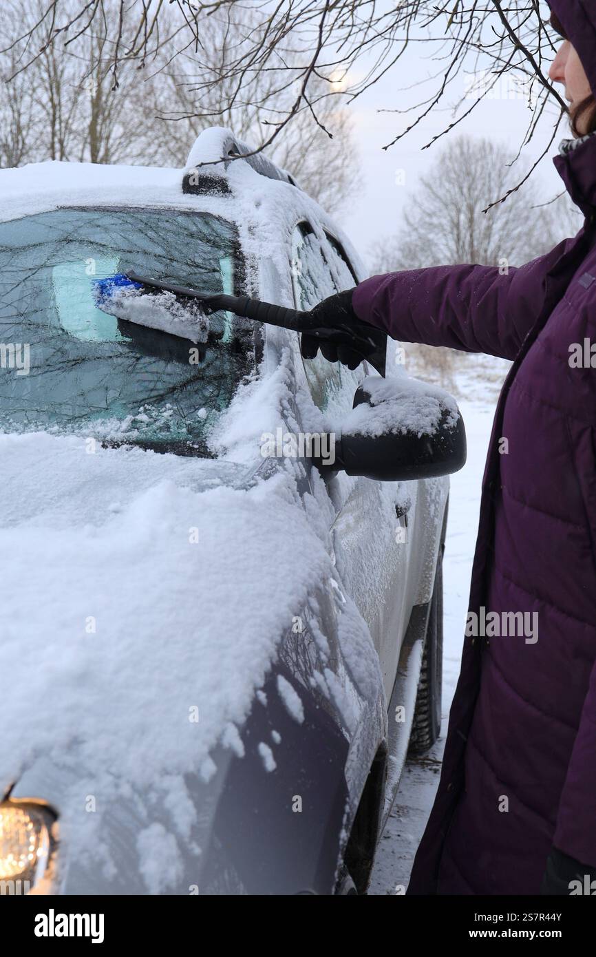 La donna proprietaria di un'auto sta in piedi lateralmente mentre pulisce la sua auto dalla neve caduta durante la notte utilizzando uno speciale orientamento verticale della spazzola Foto Stock