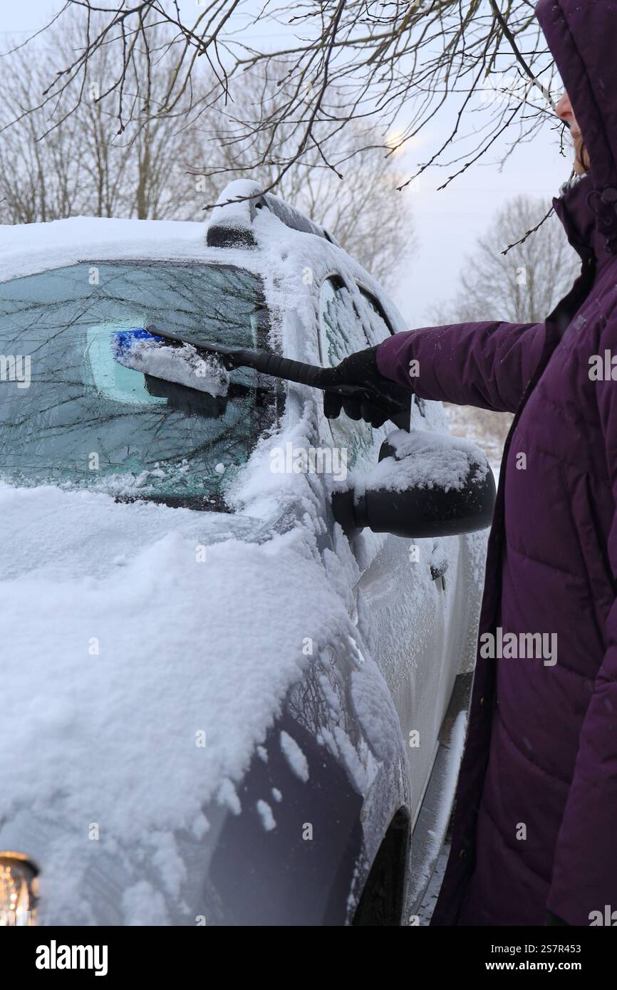 Donna in giacca calda viola con cappuccio rimuove la neve dall'auto con la spazzola per auto. Vista laterale dal basso Foto Stock
