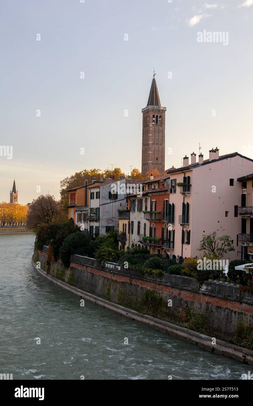 Splendida vista sul fiume di Verona, Italia, con colorati edifici storici lungo il fiume Adige, vegetazione lussureggiante e l'iconico campanile sottostante Foto Stock