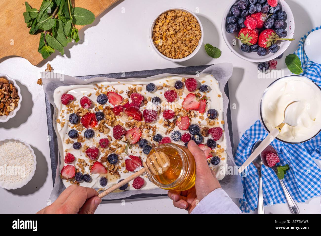 Preparazione della cottura con sfondo di corteccia di yogurt congelato con yogurt greco, bacche fresche, muesli, miele, salsa al cioccolato, con vassoio da forno deco. Vista dall'alto della donna Foto Stock