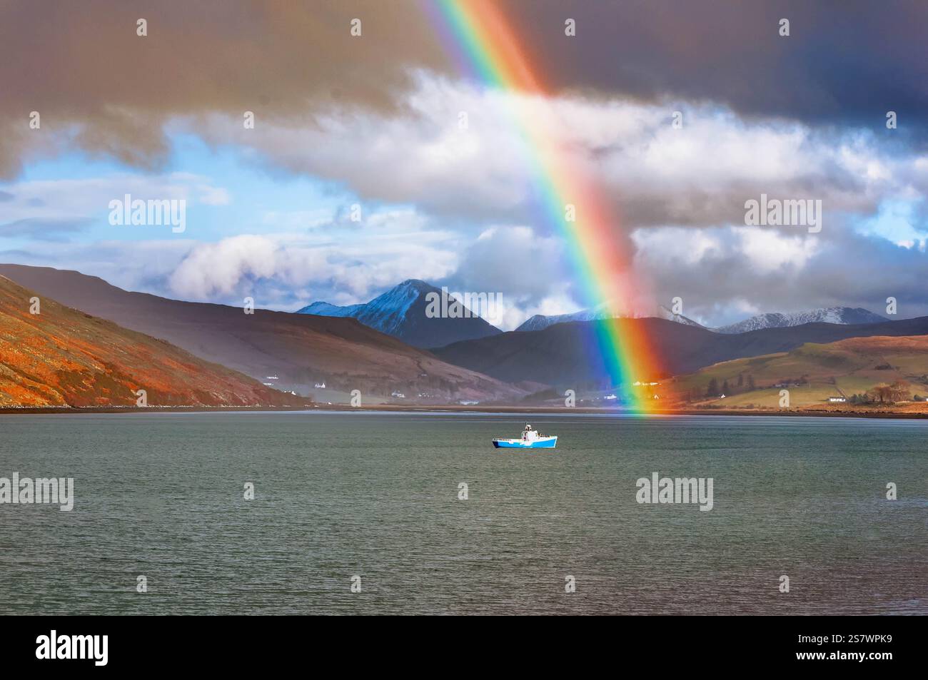 Una barca da pesca solitaria galleggia pacificamente su un mare calmo, sotto un vivace arcobaleno che si innalza su un paesaggio montuoso. La scena suggerisce un momento sereno Foto Stock