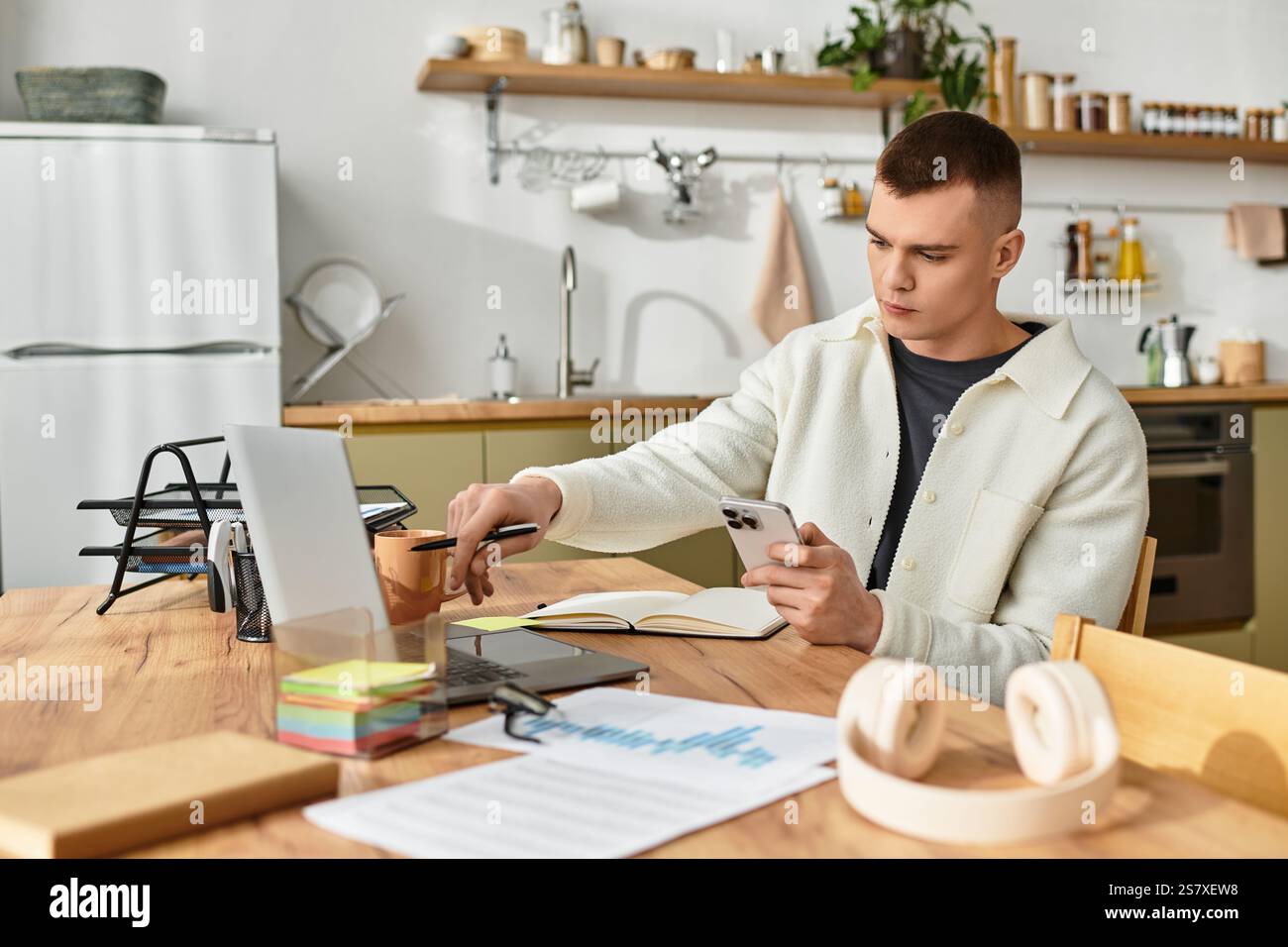 Un giovane attento si impegna a lavorare a distanza dalla sua cucina moderna, bilanciando le attività sui dispositivi. Foto Stock