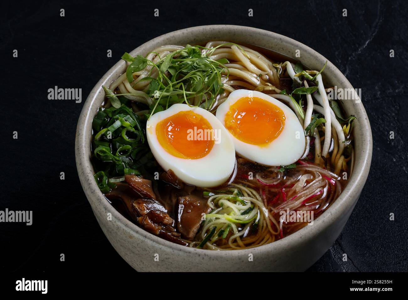 Una ciotola di ramen con uova sode perfettamente cotte, cipolle verdi vivaci, microverdure fresche, germogli di soia, spaghetti, e un ricco brodo scuro su un bl Foto Stock