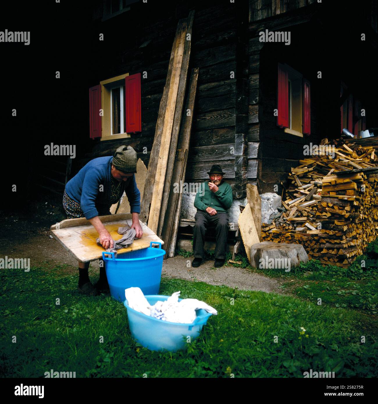 Ältere Frau wäscht Wäsche in einem blauen Becken vor einem Holzhaus, während ein rauchender Mann auf einer Bank sitzt, in Friaul-Julisch Venezien, Italien um 1976. Foto Stock