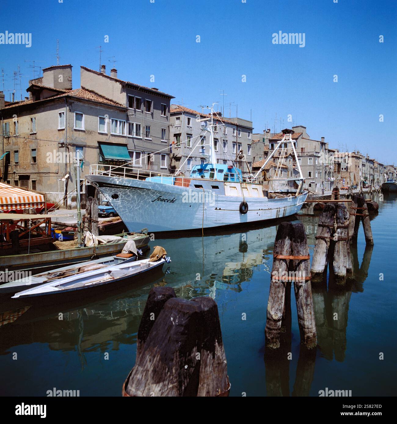Traditioneller Fischtrawler, der in Venedig, Italien, festgemacht Hat und sich an einem sonnigen Sommertag auf einer ruhigen Wasseroberfläche mit farbenfrohen Gebäuden im Hintergrund spiegelt, Italien um 1976. Foto Stock