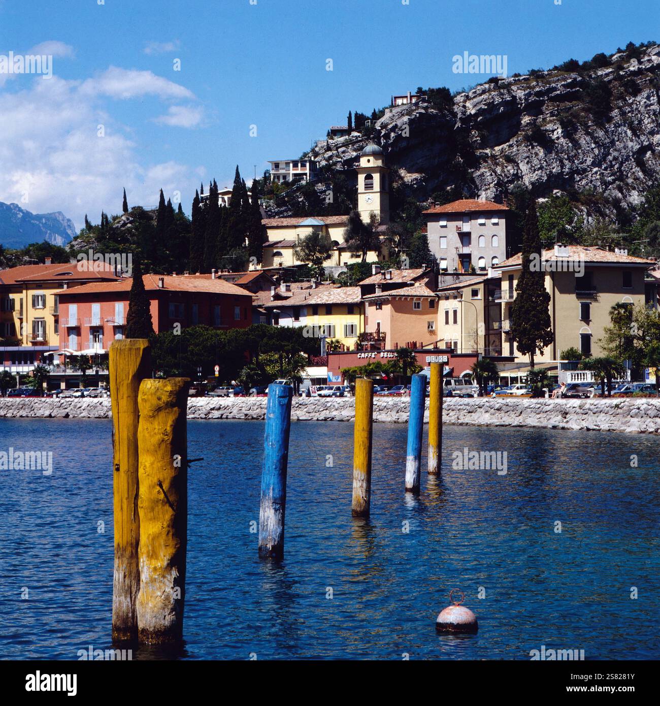 Malerische Aussicht auf die farbenfrohen Ankerpfähle, die aus dem Gardasee ragen, mit der bezaubernden Stadt Torbole am Gardasee, die sich an den Berghang schmiegt, Italien um 1977. Foto Stock