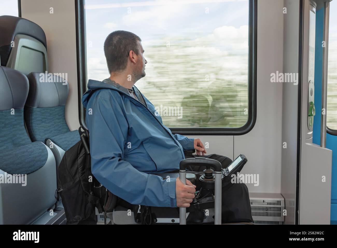 Viaggiatore di sesso maschile con disabilità che si gode la vacanza e il viaggio in treno in Europa, seduto su una sedia a rotelle che guarda fuori dal finestrino. Foto Stock