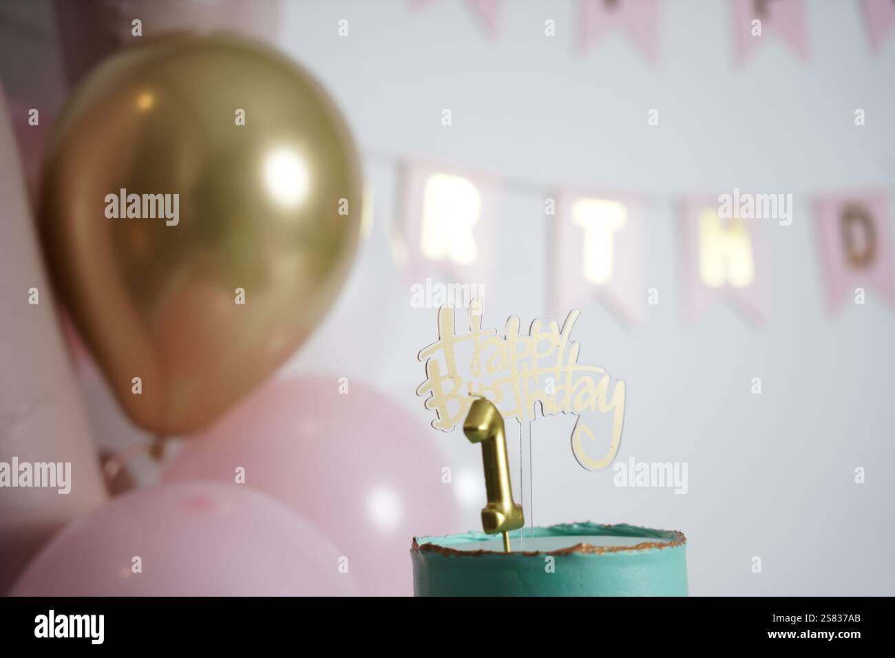 Una torta di compleanno color tè con accenti dorati, condita con un condimento dorato "Happy Birthday" e una candela numero 1. Lo sfondo sfocato presenta una spilla pastello Foto Stock