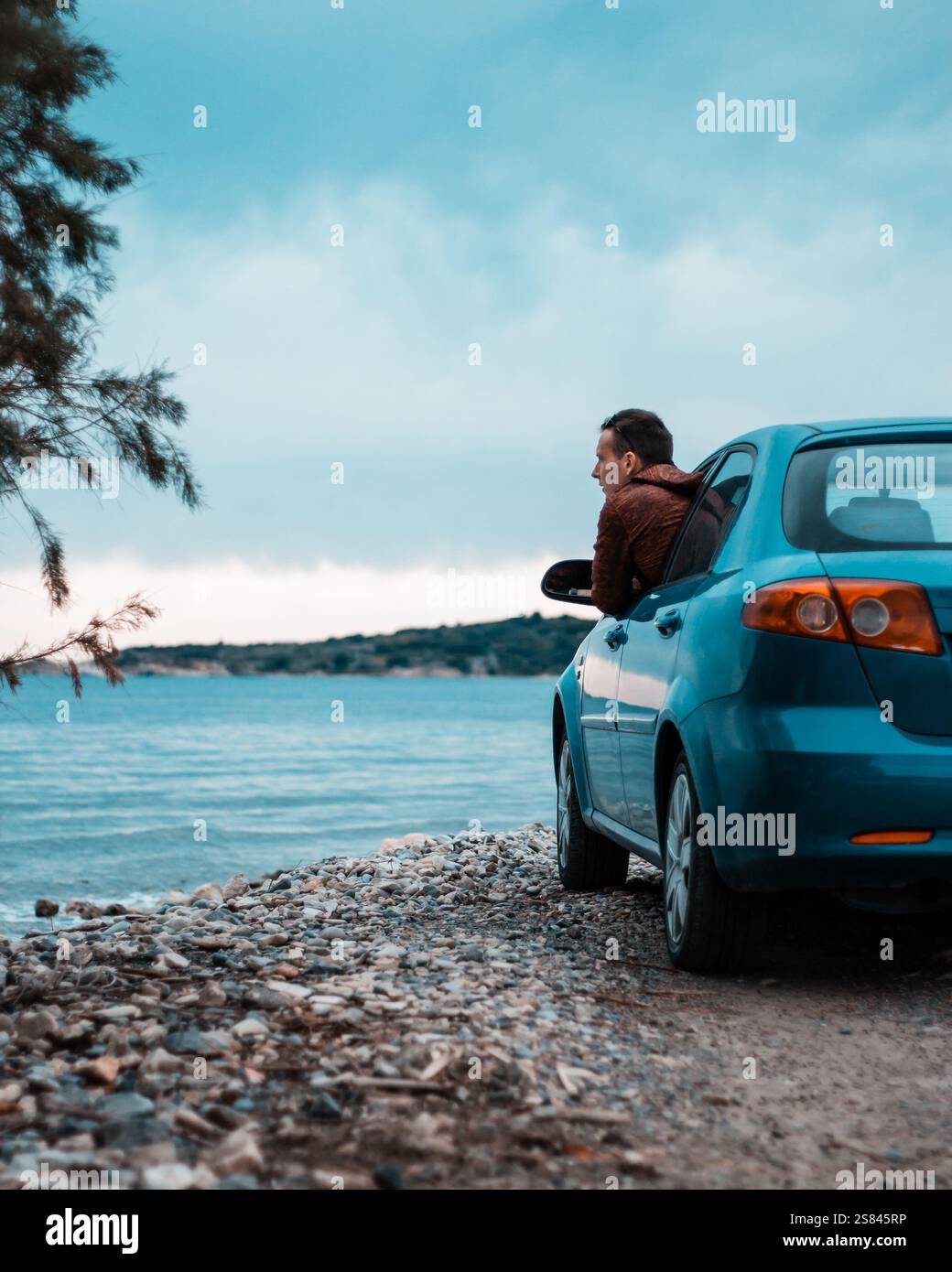 Un uomo si appoggia su un'auto blu vicino a una costa rocciosa, di fronte a un grande specchio d'acqua. Un ramo d'albero incornicia la scena, con colline lontane sotto cieli nuvolosi. Foto Stock
