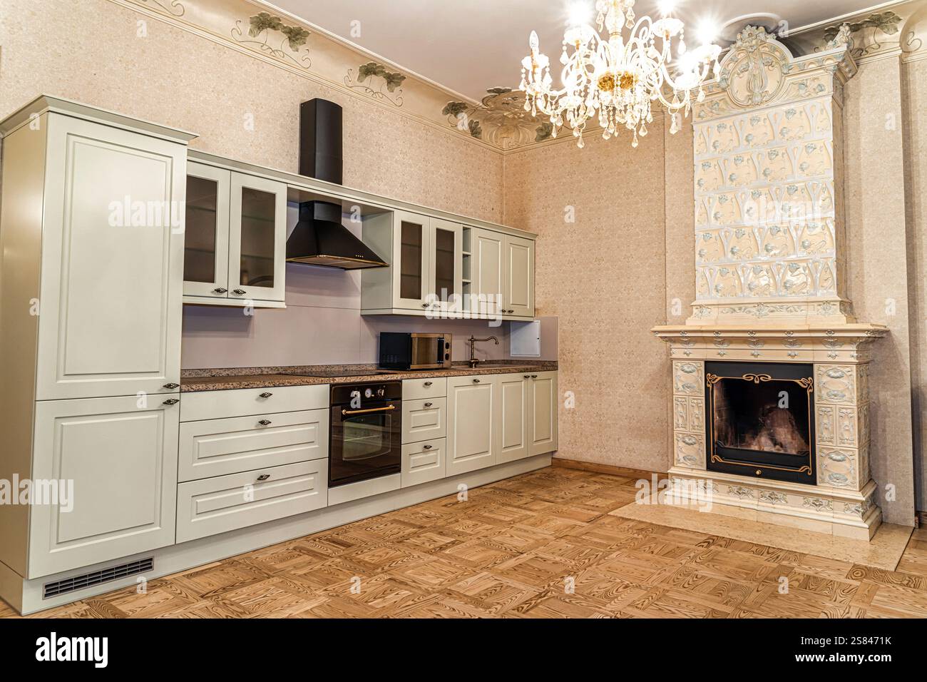 Armadietti luminosi con porte in vetro, cappa nera, caminetto decorato con piastrelle, lampadario di cristallo e pavimento in legno geometrico lucido. Foto Stock