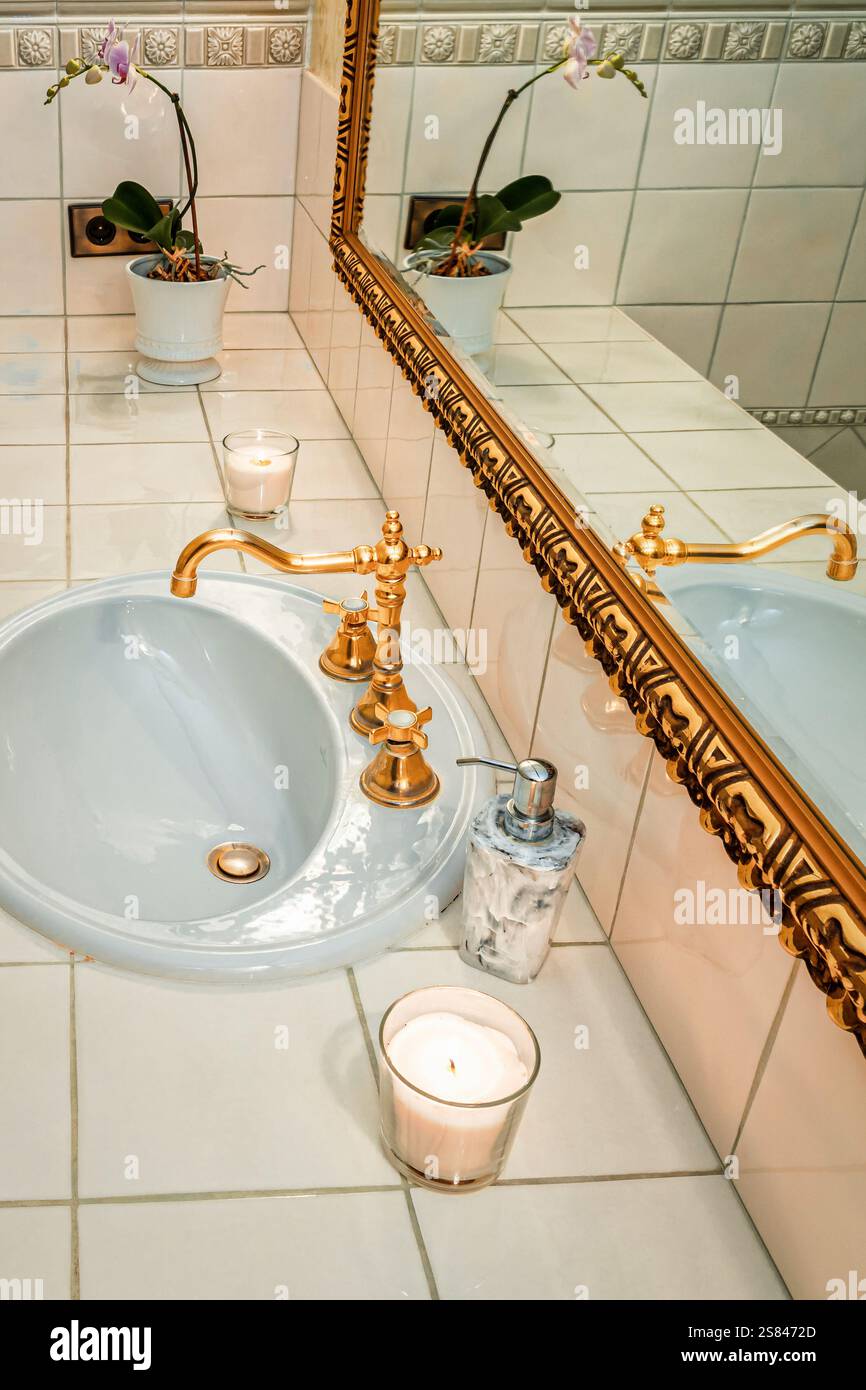 Lavello in ceramica bianca con rubinetto dorato ornato, candele accese e un'orchidea in vaso con fiori viola su un piano di appoggio piastrellato vicino a un grande specchio incorniciato in oro Foto Stock