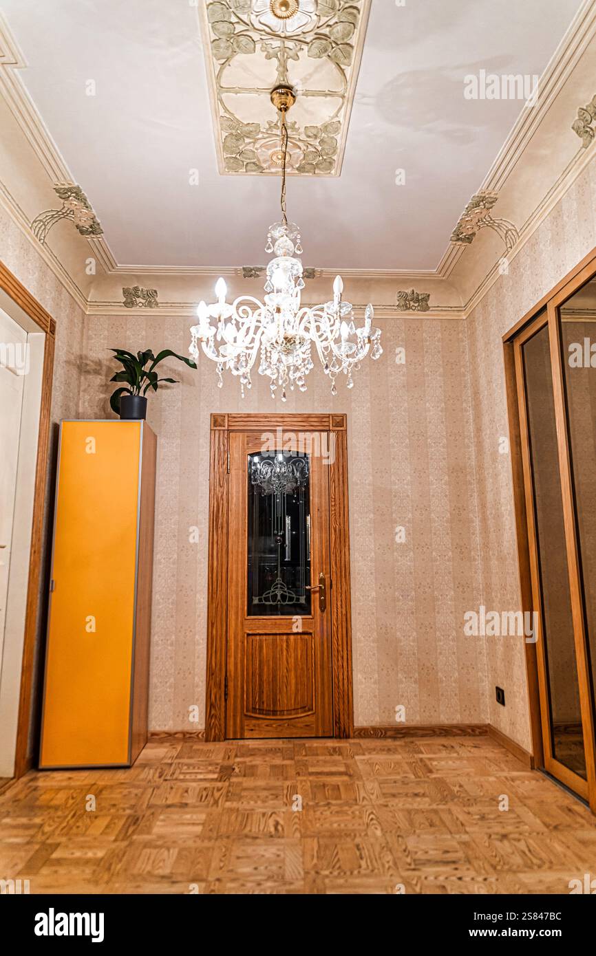 Spazio interno caratterizzato da una porta in legno con pannello di vetro, lampadario di cristallo, soffitto in gesso floreale, carta da parati a motivi geometrici e pavimento in parquet. Foto Stock