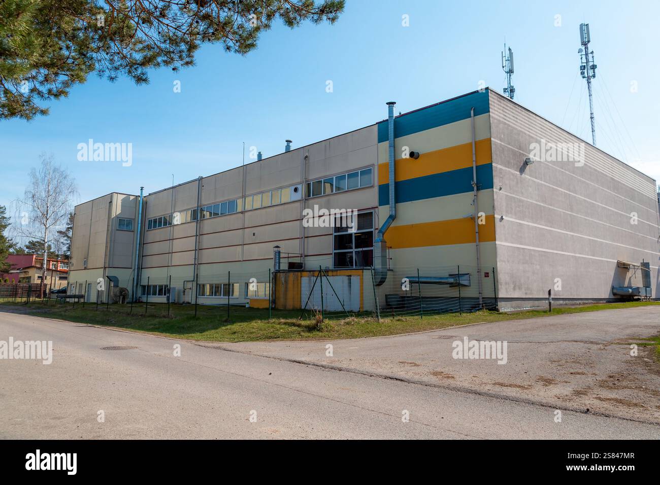 Grande edificio di stile industriale con strisce gialle e blu, area recintata, installazioni di servizi e torri cellulari sul tetto sotto un cielo limpido. Foto Stock