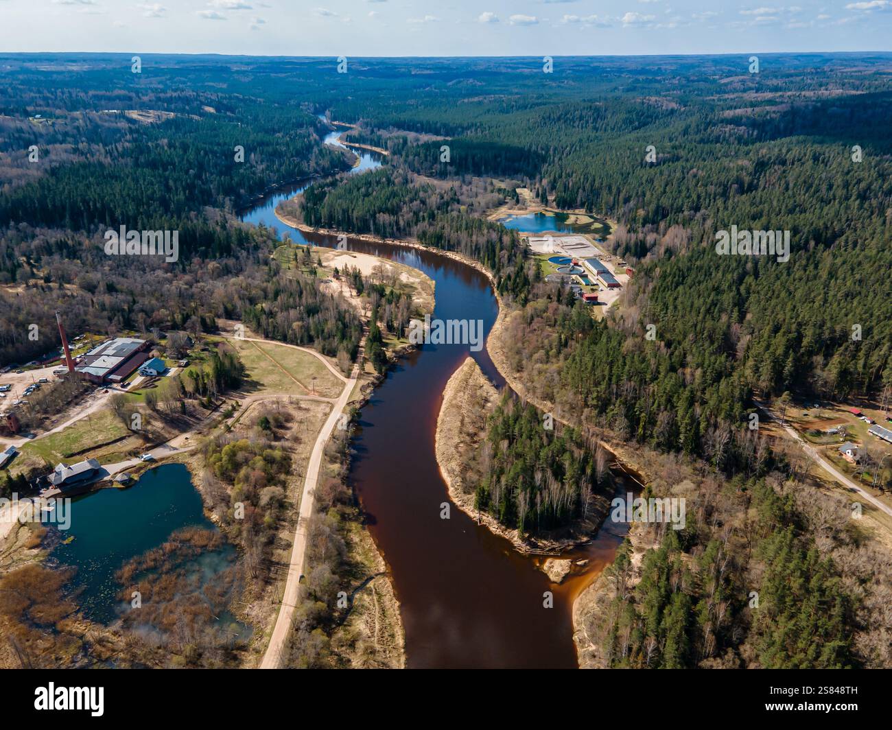 Prospettiva aerea di un fiume che si snoda tra foreste e colline, con edifici industriali sulla sinistra e strutture colorate sulla destra. Foto Stock