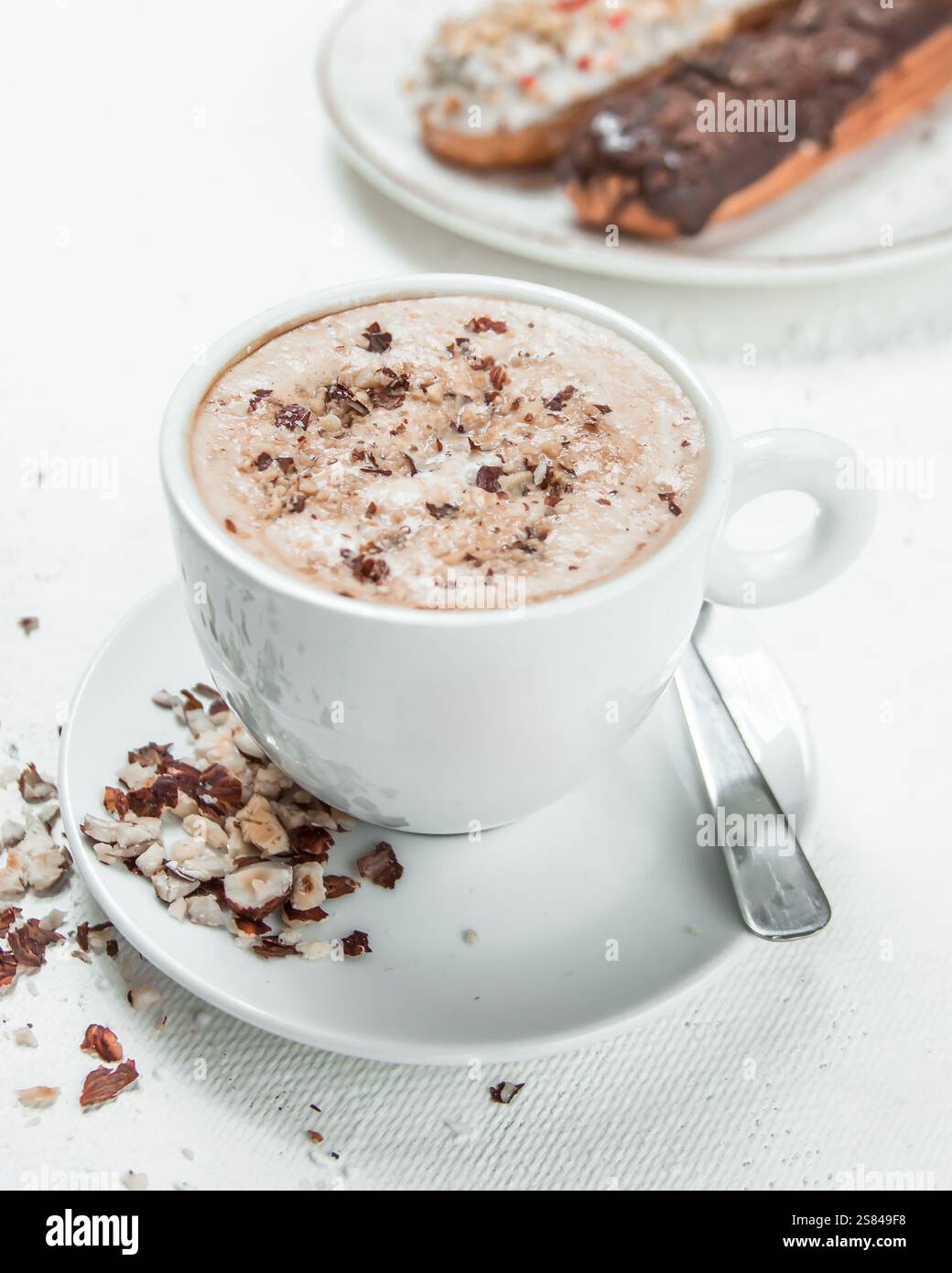 Una tazza di caffè bianco con una bevanda frizzante e condimenti schiacciati su un piattino, accompagnata da eclari con cioccolato e glassa spolverata su una superficie bianca. Foto Stock
