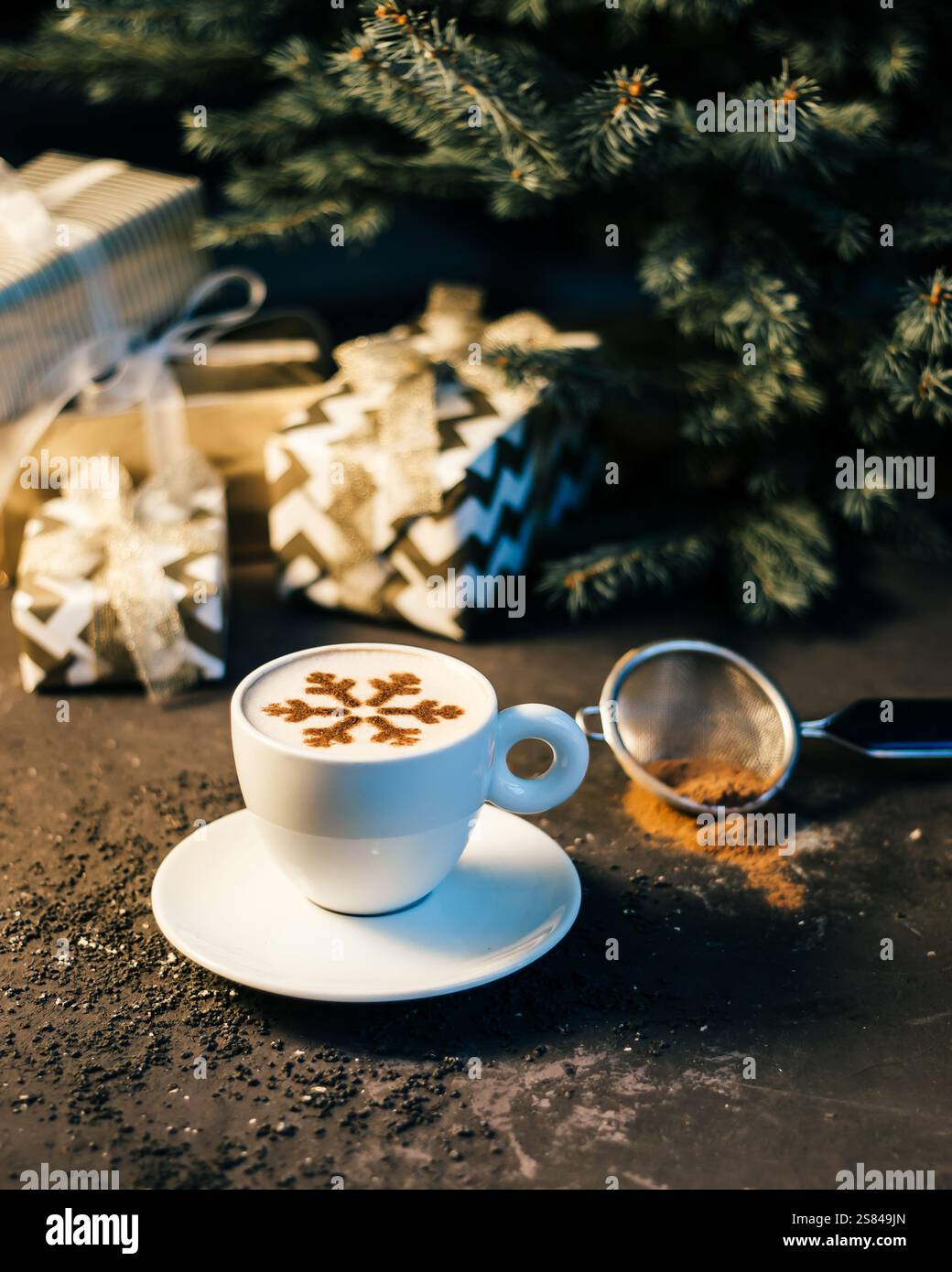 Una tazza di caffè bianca con un motivo di cacao con fiocco di neve sulla schiuma, circondata da regali avvolti, un setacciatore di cacao e un ramo di abete su una superficie scura. Foto Stock