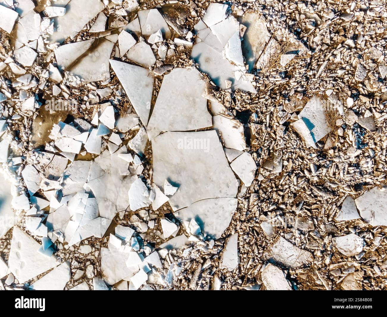 Spaccature e rotture di ghiaccio di varie dimensioni sparse su una superficie ruvida con piccoli frammenti di ghiaia e tonalità terrose sullo sfondo Foto Stock