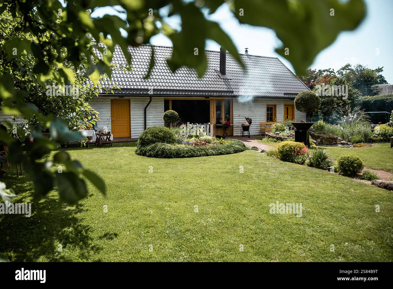Un gruppo si riunisce vicino a un barbecue su un patio in un cortile con erba rifilata, cespugli paesaggistici, un sentiero da giardino e alberi sospesi. Foto Stock