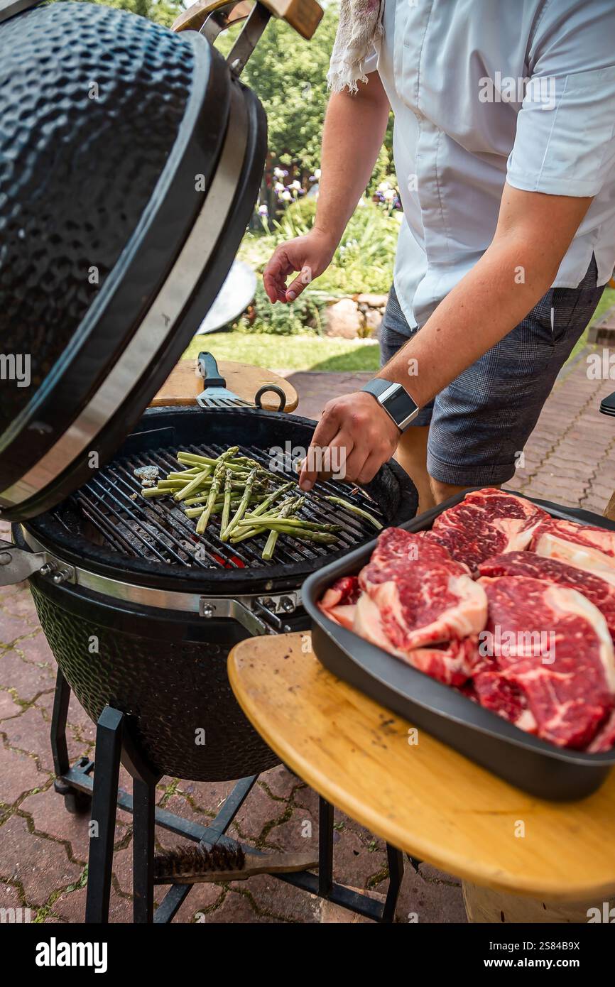 Una persona grigia gli asparagi su una griglia in ceramica nera con coperchio a cupola, mentre le bistecche di marmo crude riposano su un tavolo di legno in un patio con giardino. Foto Stock