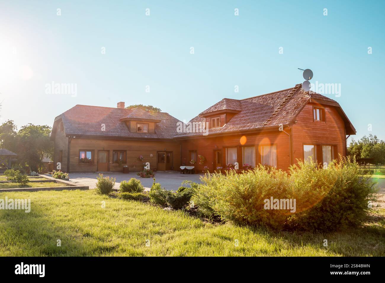 Grande casa in legno con tetto inclinato e parabola satellitare, circondata da un prato ben tenuto, cespugli e alberi sotto la calda luce del sole in un setti rurale Foto Stock