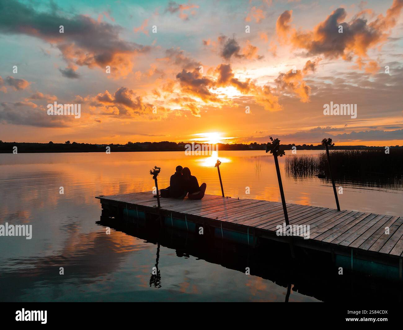 Due persone siedono su un molo di legno che si estende su un lago calmo, caratterizzato da un vibrante tramonto con sfumature arancioni e gialle. Reeds e linea ad albero visibili. Foto Stock