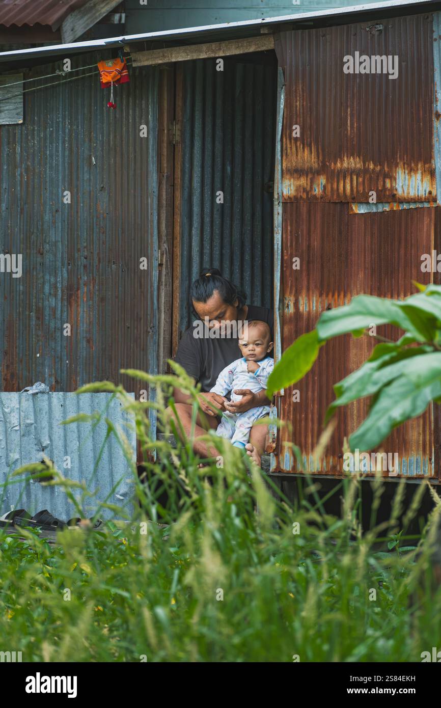 Balikpapan, Indonesia - 25 dicembre 2024. Il giovane padre culla dolcemente il suo bambino tra le braccia, fissandolo davanti al loro ferro ondulato Foto Stock