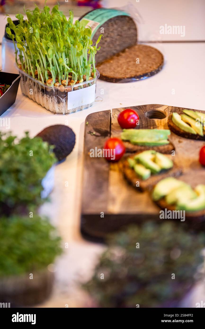 Fette di pane scuro con avocado e pomodori ciliegini su un tagliere, circondate da germogli di piselli, vegetazione e una pagnotta di pane su una superficie bianca. Foto Stock