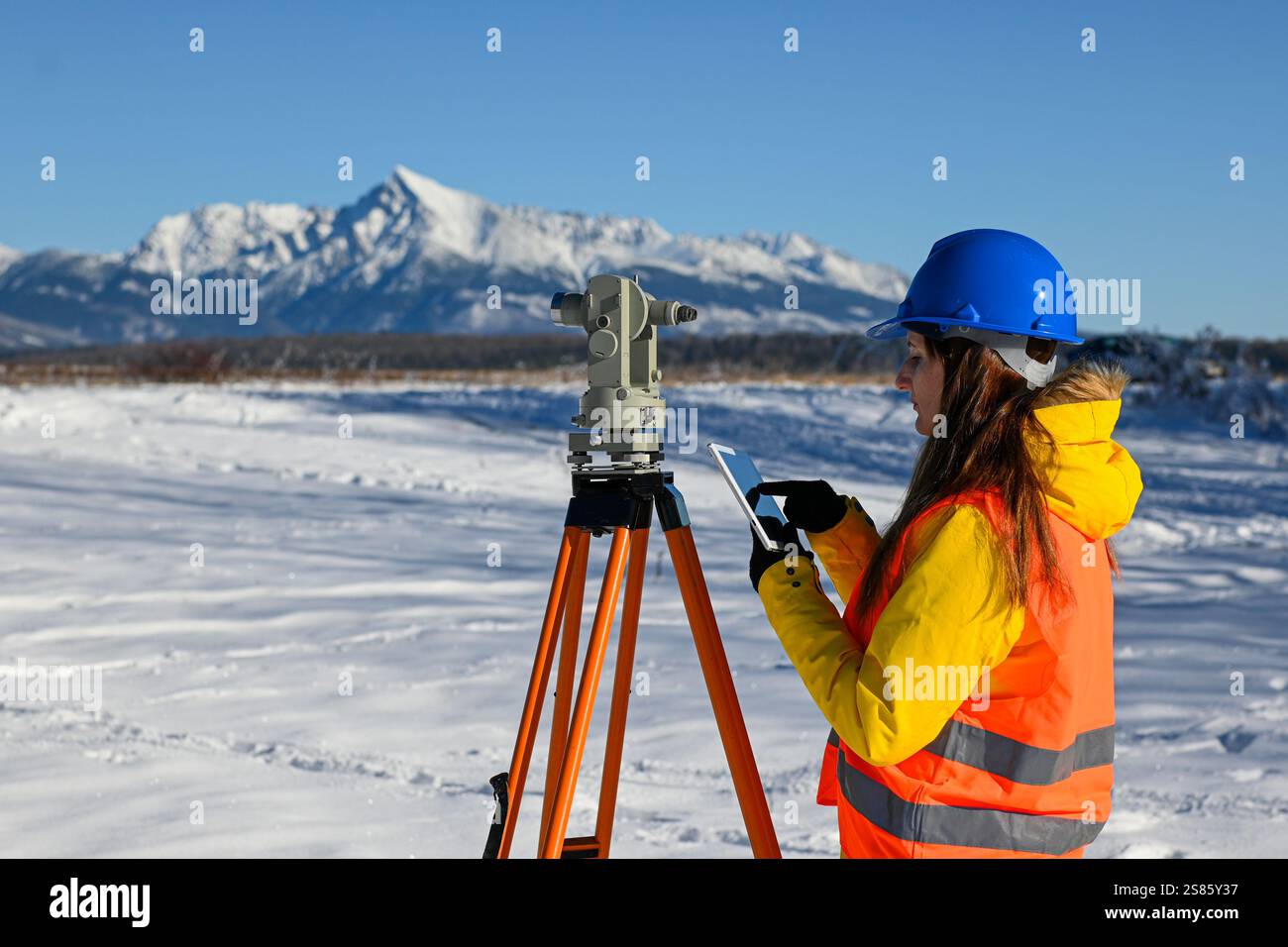 Topografia femminile che utilizza teodolite e tablet computer per la topografia in paesaggi innevati di montagna. Foto Stock