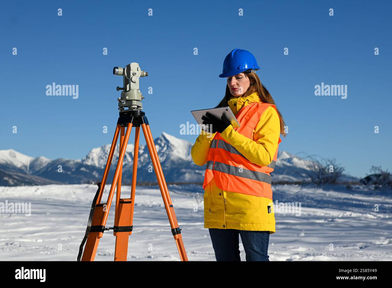 Topografia femminile che utilizza teodolite e tablet computer per la topografia in paesaggi innevati di montagna. Foto Stock