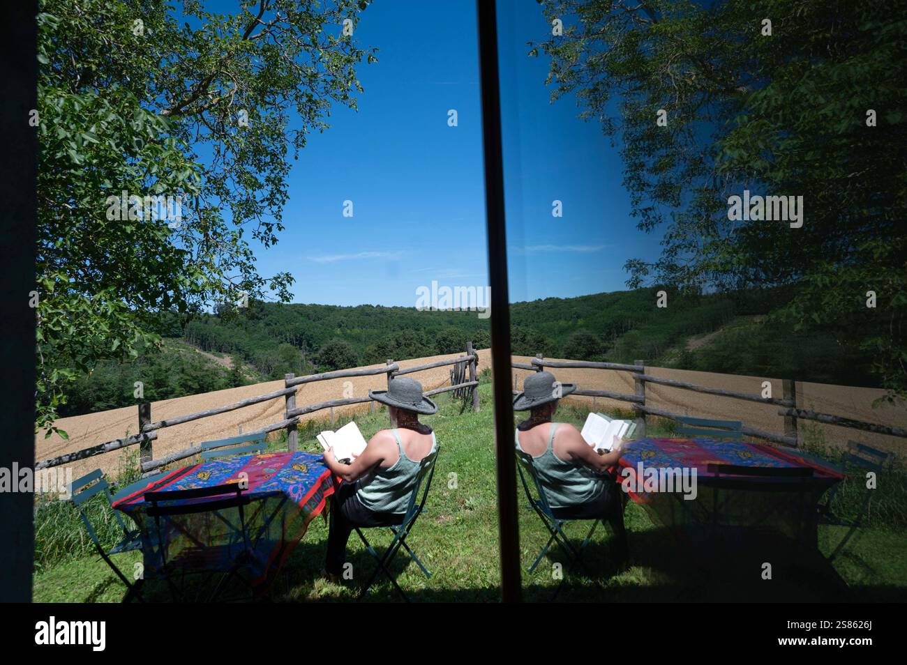 Vacanze estive e lettura: Donna seduta sulla sedia fuori a leggere un romanzo al sole estivo. Riflesso in una finestra e vista sulla campagna Foto Stock
