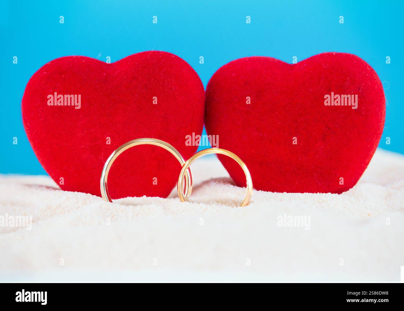 Due cuori di velluto rosso su uno sfondo di sabbia con un cielo blu, accompagnati da fedi nuziali dorate. Perfetto per i temi di San Valentino, i matrimoni Foto Stock