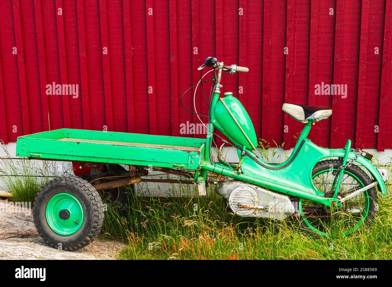 Un vivace ciclomotore verde a tre ruote con alette è parcheggiato accanto ad un rustico edificio in legno rosso. Il ciclomotore si appoggia su un'erba, mostrando il suo dist Foto Stock