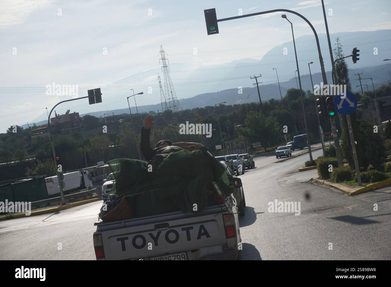 Dopo la raccolta delle olive un lavoratore stanco sdraiato su sacchi di olive nel retro di un camion in movimento guarda la telecamera e alza la mano nel saluto Foto Stock