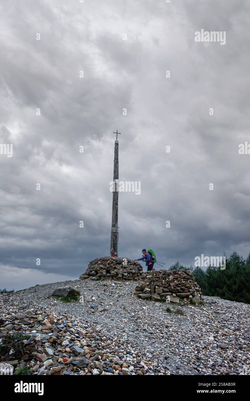 Pellegrino sul cammino di Santiago che offre pietre votive e offerte in Cruz de ferro (Croce di ferro), collina di Foncebadón, regione di Bierzo, Castiglia e León, Spagna. Foto Stock