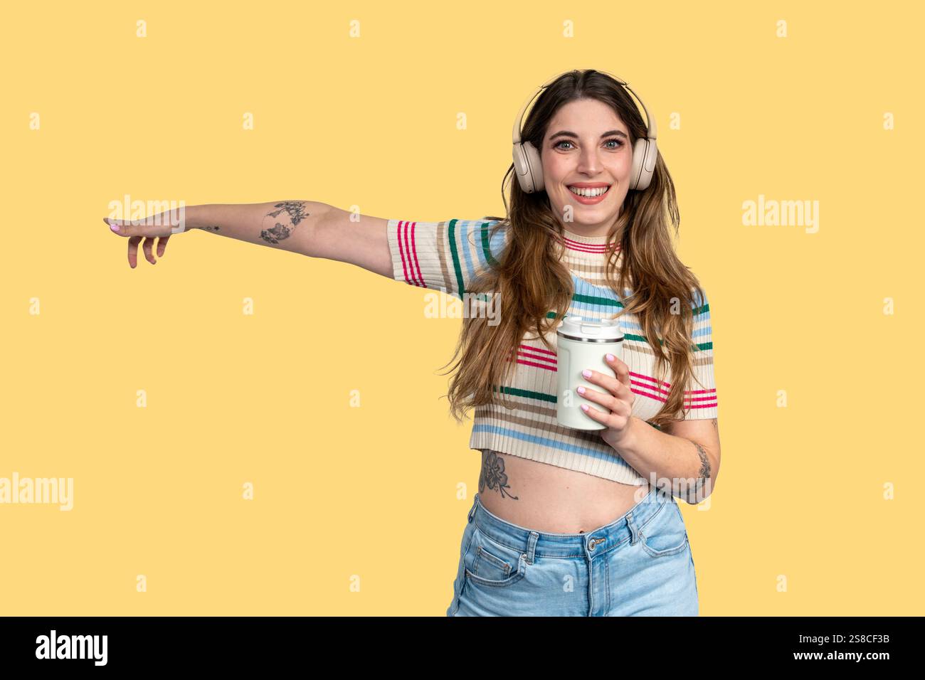 Giovane donna sorridente con tatuaggi che punta il dito e tiene in mano una tazza da caffè riutilizzabile mentre ascolta musica con le cuffie su sfondo giallo Foto Stock