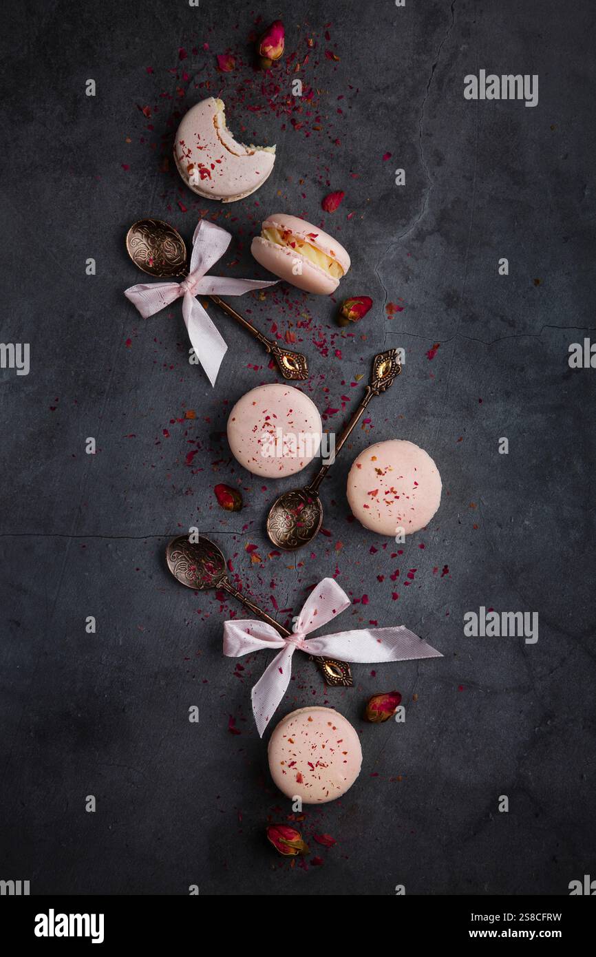 Macaron rosa decorati con petali di rosa essiccati, con cucchiai vintage con nastri, disposti su una superficie scura per una presentazione elegante Foto Stock