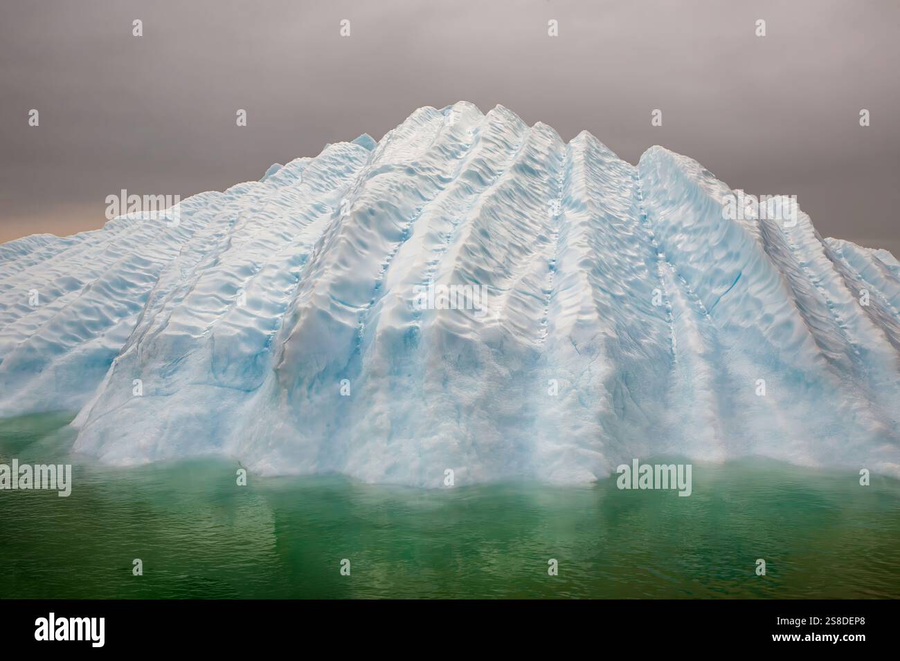 Un enorme iceberg con creste impressionanti e una vivace tonalità blu è visto galleggiare in acque verdi calme. Il cielo nuvoloso crea uno sfondo drammatico, hinti Foto Stock
