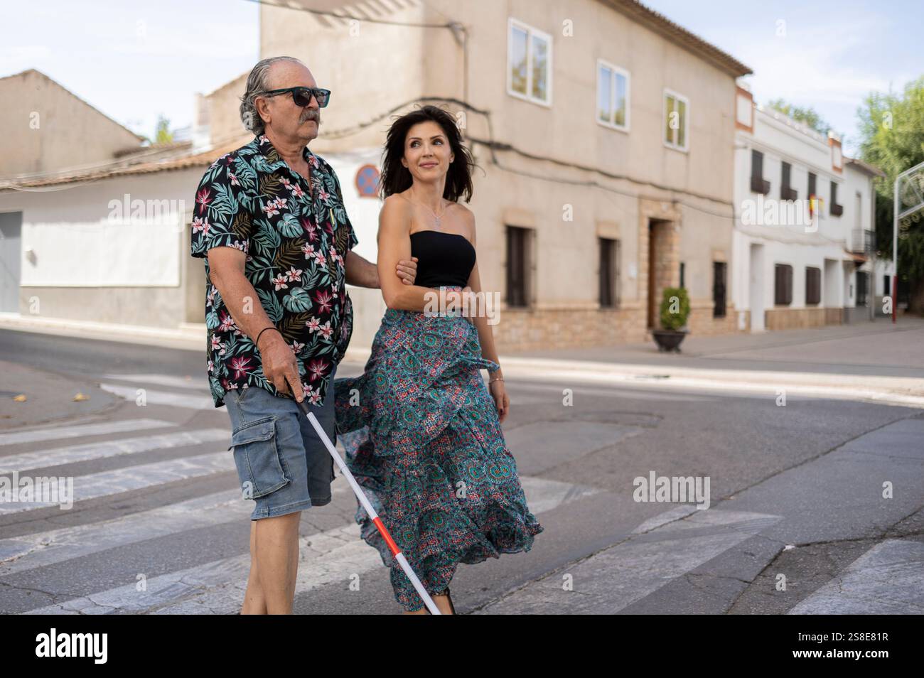 Un padre cieco usa un bastone bianco mentre sua figlia lo guida attraverso una tranquilla strada cittadina. Entrambi indossati con un abbigliamento casual estivo, sono perfetti da sfoggiare Foto Stock