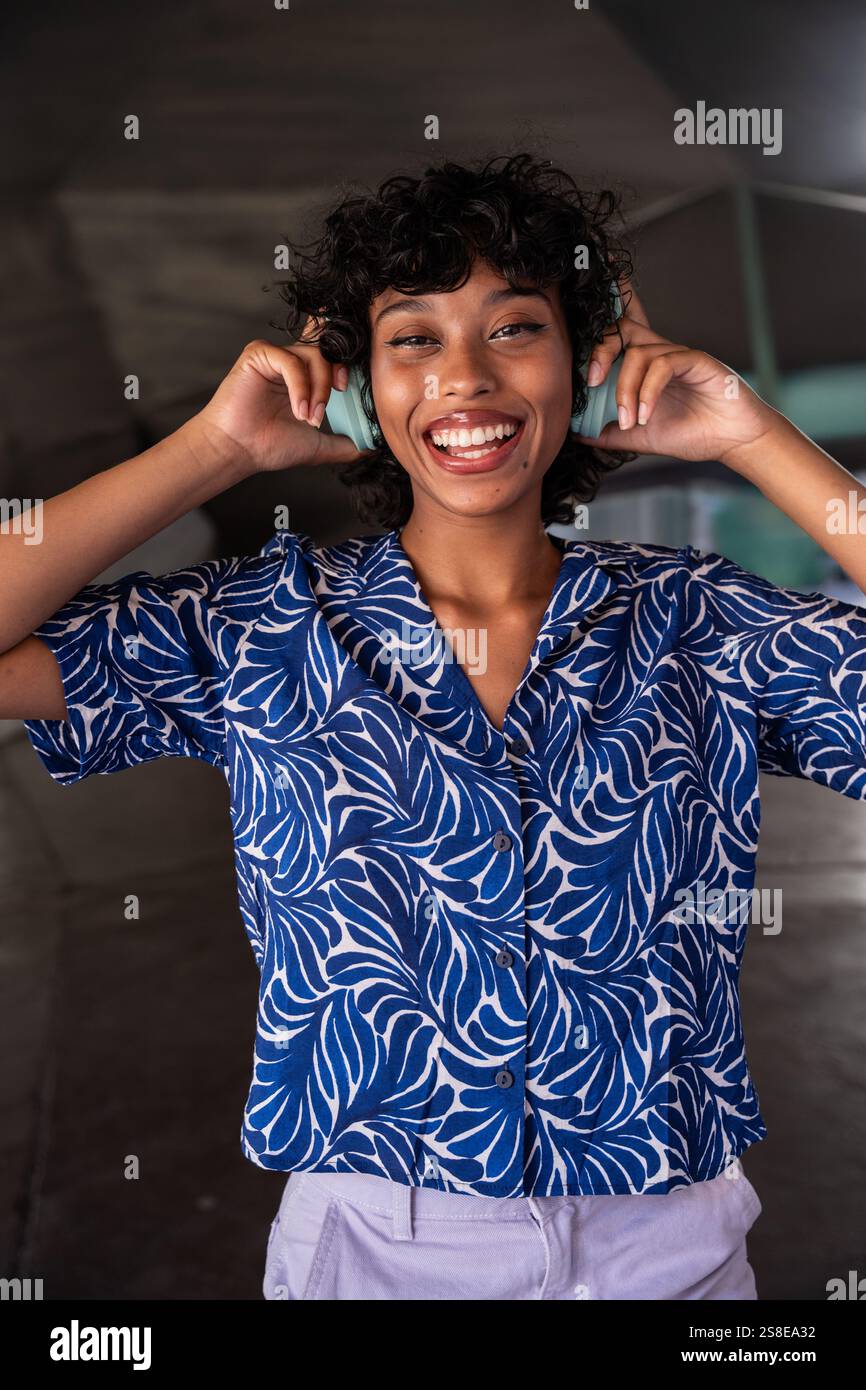 Una giovane donna allegra che indossa una camicia colorata blu vivace e ascolta con gioia la musica con le cuffie. Il suo ampio sorriso e gli occhi espressivi trasmettono Foto Stock