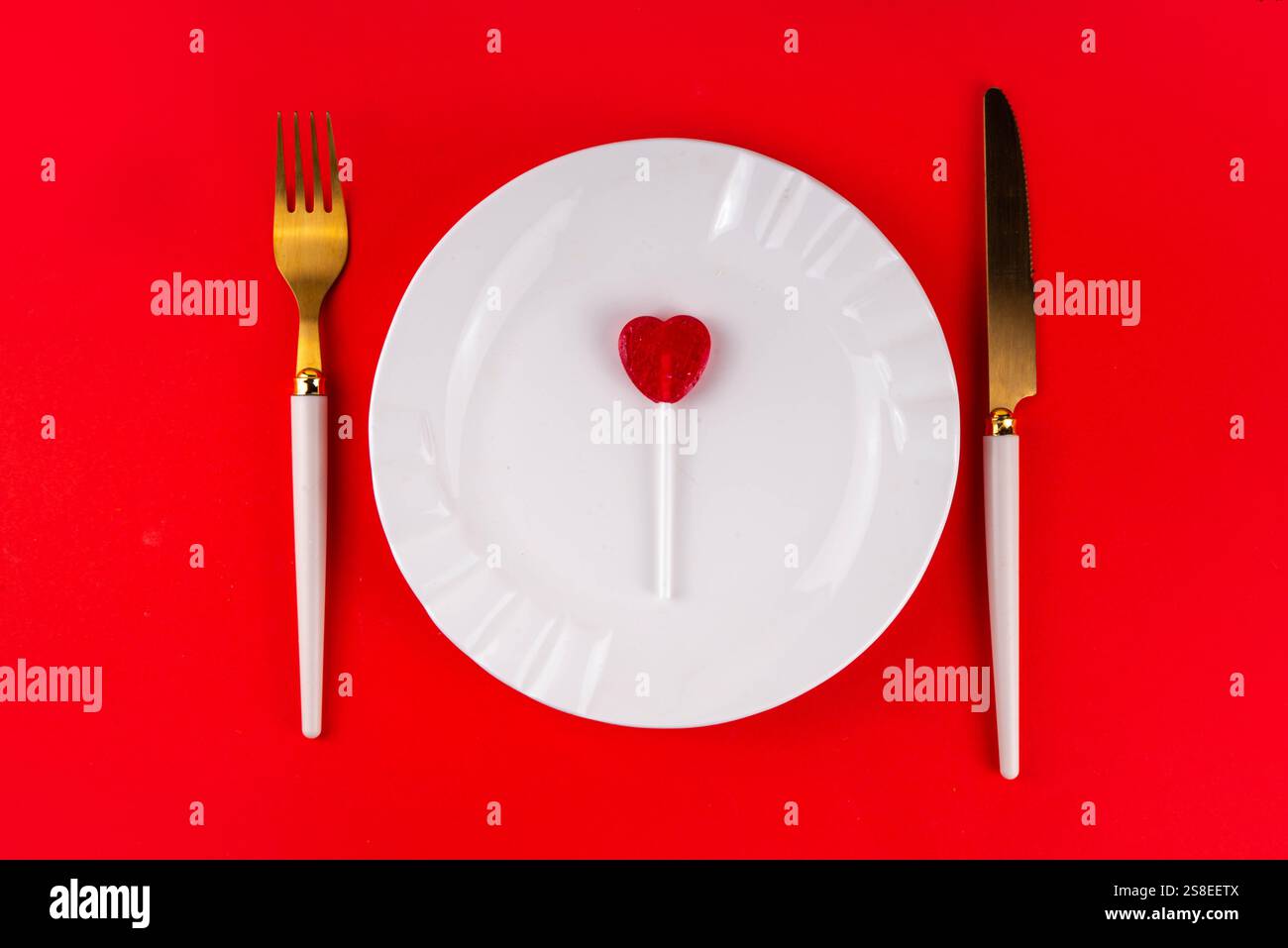 Posate di San Valentino con piatto, coltello, forchetta e lollypop a forma di cuore di zucchero rosso, su sfondo rosso brillante, vista dall'alto spazio di copia. Festività, va Foto Stock