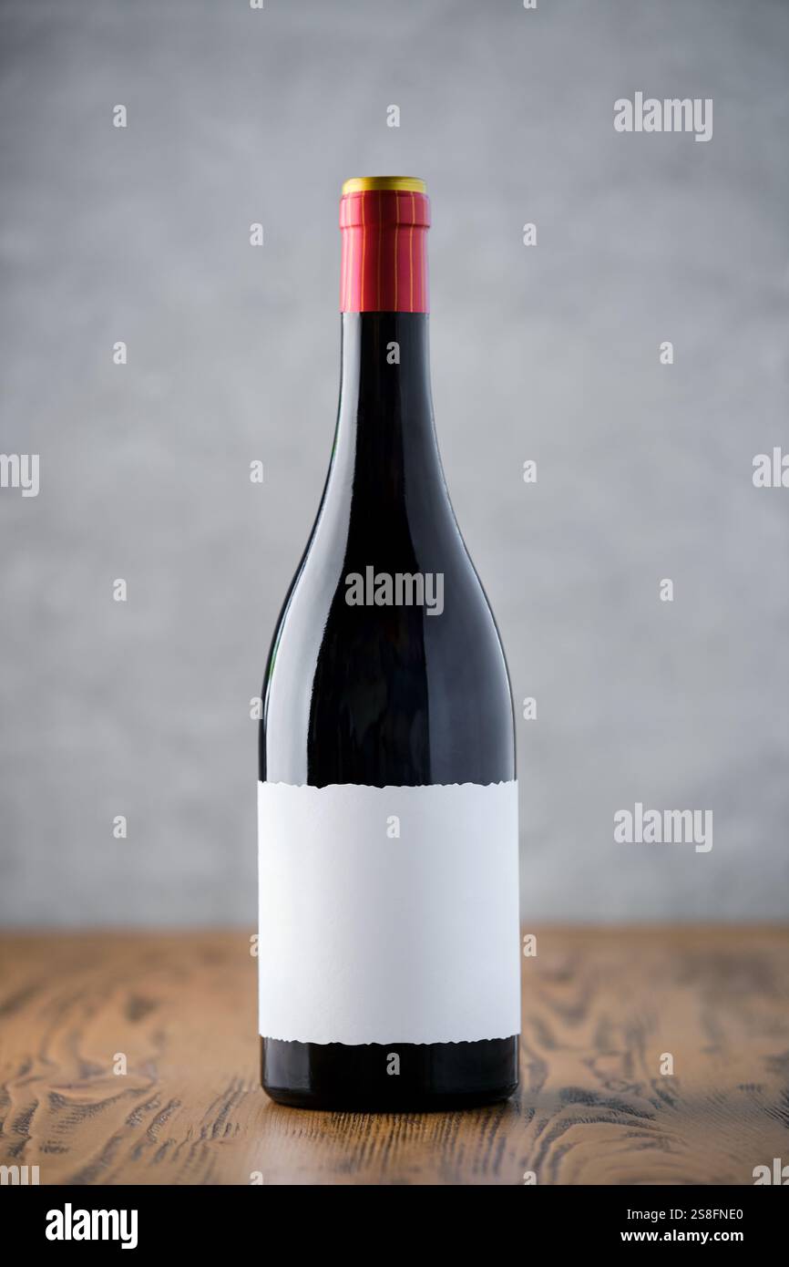 Una singola bottiglia di vino scuro con un'etichetta vuota si staglia su una superficie in legno, su un morbido sfondo grigio, pronta per essere marchiata. Foto Stock