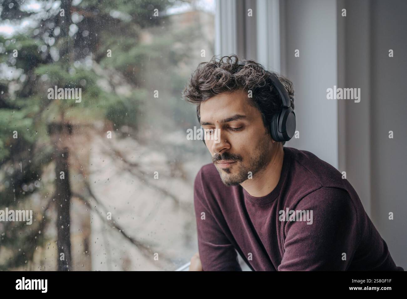 Bell'uomo mediorientale con cuffie sovrauricolari che ascoltano musica e si rilassano vicino alla finestra. Foto Stock