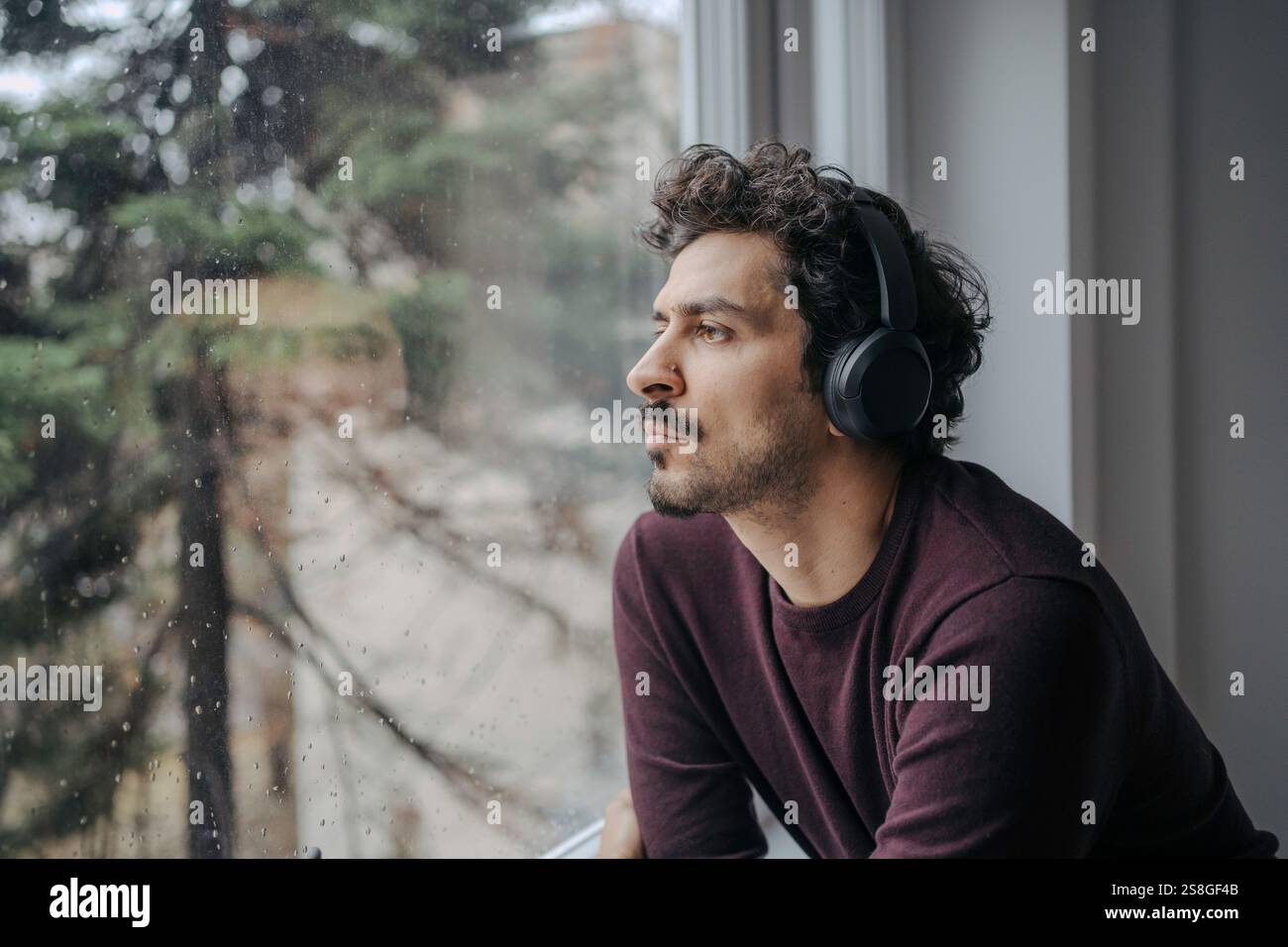 Bell'uomo mediorientale con cuffie sovrauricolari che ascoltano musica e si rilassano vicino alla finestra. Foto Stock